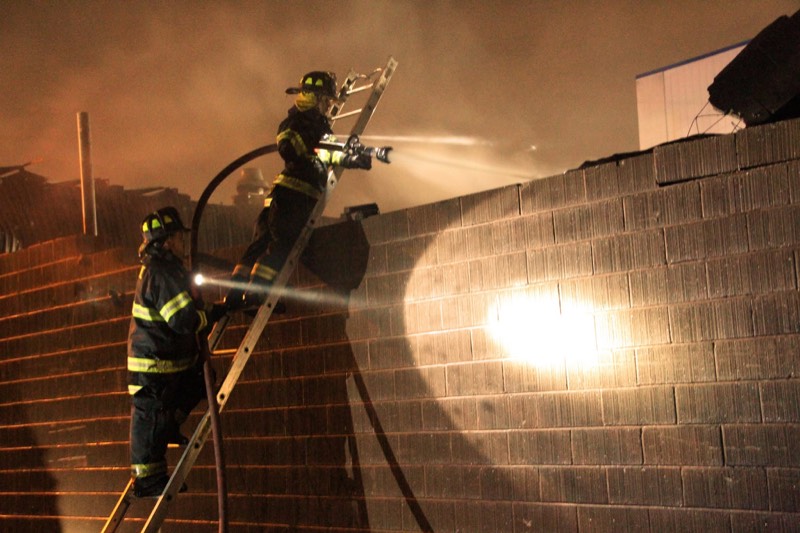 Two firefighters on a ladder against a block wall, one aiming a hose stream into the smoke at night