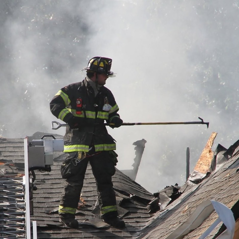 A firefighter standing on a roof with a hook as smoke rises around him