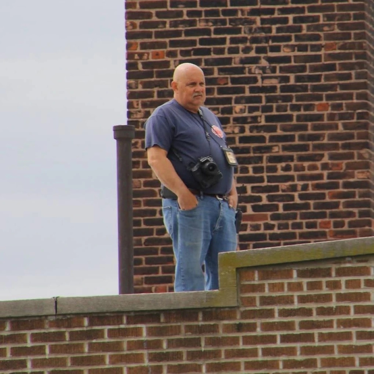 Ron Johnson standing on a rooftop beside a brick chimney with his camera around his neck