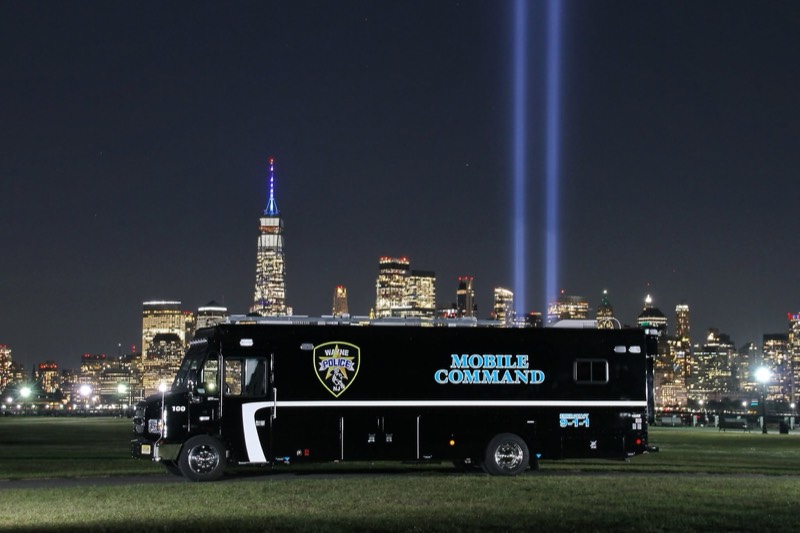 A black police mobile command vehicle on the lawn at night with the Lower Manhattan skyline and Tribute in Light beams behind it