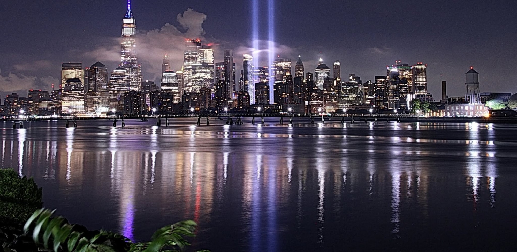 The twin beams of the Tribute in Light rising over the Lower Manhattan skyline, reflected in the Hudson River at night